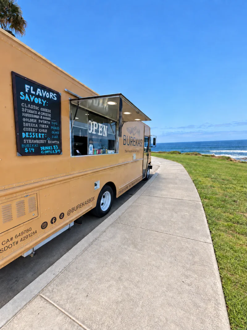 The caramel-colored Burekas Box electric food truck beside the ocean with its service window open
