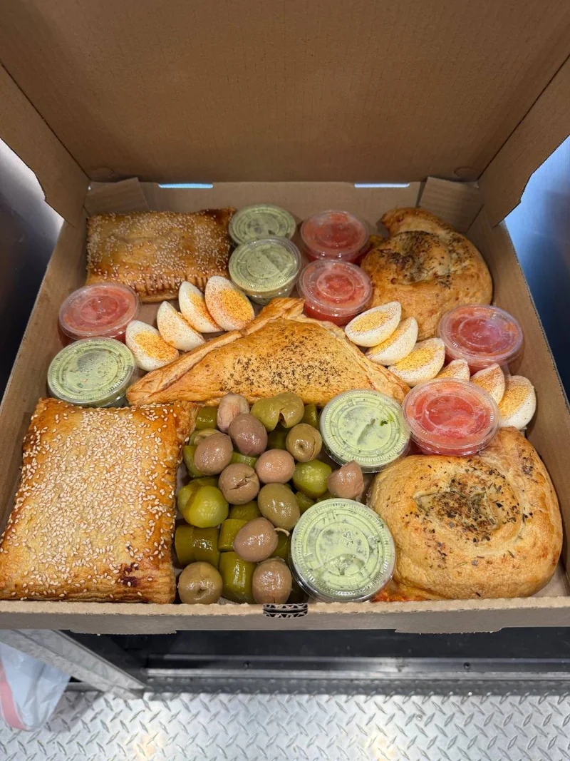 An open Burekas Box packed with sesame-topped and spiral burekas, seasoned eggs, olives, pickles, grated tomato and BBox Green Sauce